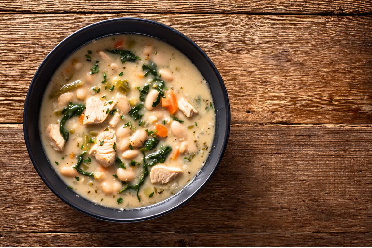 Overhead view of High Protein Creamy Garlic Chicken Soup in a matte black bowl on a rustic wooden table. Creamy broth filled with chunks of chicken breast, white beans, diced carrots, spinach, celery, and fresh herbs.