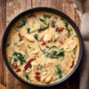 Overhead view of creamy Tuscan chicken soup in a black ceramic bowl on a rustic wooden table. Golden cream broth surrounds chunks of chicken, white beans, fresh spinach, and red sun-dried tomatoes. Beige linen napkin beside the bowl.