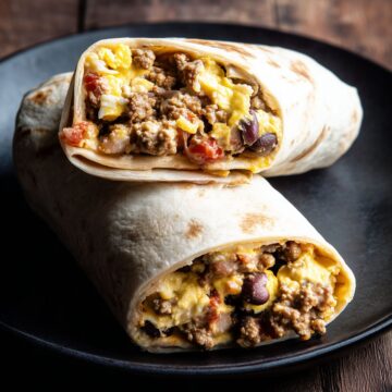 Two High Protein Taco Breakfast Burrito halves stacked on a matte black plate. The lightly browned flour tortillas reveal a filling of seasoned ground turkey, scrambled eggs, melted cheese, black beans, and salsa against a dark rustic wooden background.