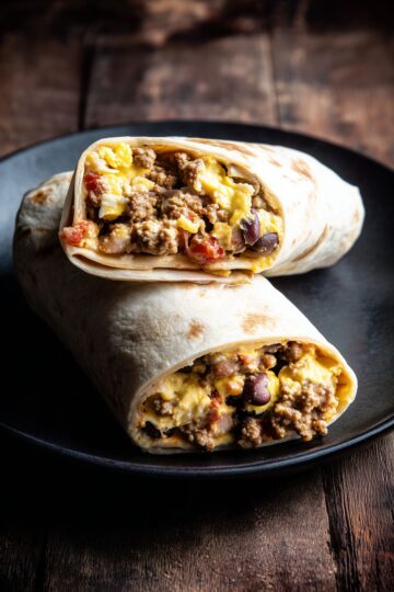 Two High Protein Taco Breakfast Burrito halves stacked on a matte black plate. The lightly browned flour tortillas reveal a filling of seasoned ground turkey, scrambled eggs, melted cheese, black beans, and salsa against a dark rustic wooden background.