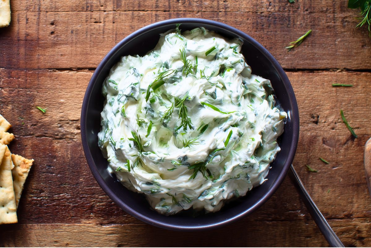 Overhead view of Holiday Herb Cream Cheese Dip in a black serving bowl. Thick, creamy white dip mixed with fresh parsley, chives, and dill, surrounded by crackers and scattered herbs on a rustic wooden table.