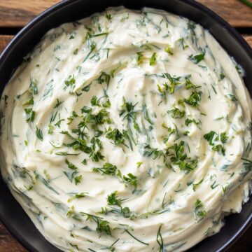 Close-up of Holiday Herb Cream Cheese Dip in a black bowl, featuring a thick, smooth, creamy texture with swirls of cream cheese and chopped fresh green herbs. Crackers and chives rest on a rustic wooden background.