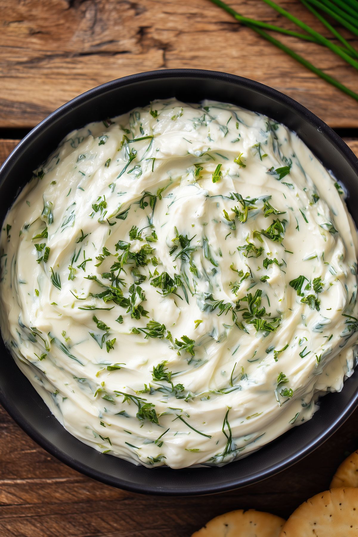 Close-up of Holiday Herb Cream Cheese Dip in a black bowl, featuring a thick, smooth, creamy texture with swirls of cream cheese and chopped fresh green herbs. Crackers and chives rest on a rustic wooden background.