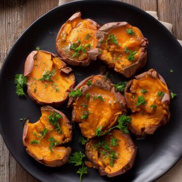 Close-up overhead view of seven honey butter smashed sweet potatoes on a matte black plate. The tender orange sweet potatoes have slightly browned edges, glossy honey butter glaze, and fresh parsley garnish. Rustic wooden table with a beige napkin underneath.