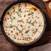 Overhead view of hot white pizza dip in a black cast iron skillet. Creamy melted cheese with golden brown spots is topped with fresh basil strips. Toasted baguette slices and a beige napkin sit on a rustic wooden table.