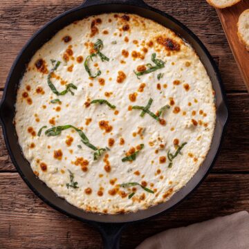 Overhead view of hot white pizza dip in a black cast iron skillet. Creamy melted cheese with golden brown spots is topped with fresh basil strips. Toasted baguette slices and a beige napkin sit on a rustic wooden table.