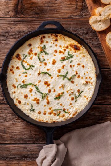 Overhead view of hot white pizza dip in a black cast iron skillet. Creamy melted cheese with golden brown spots is topped with fresh basil strips. Toasted baguette slices and a beige napkin sit on a rustic wooden table.