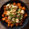 Overhead view of a Jalapeno Popper Chicken Sweet Potato Bowl with roasted sweet potato cubes, creamy seasoned chicken, melted cheese and sliced green onions in a black bowl.
