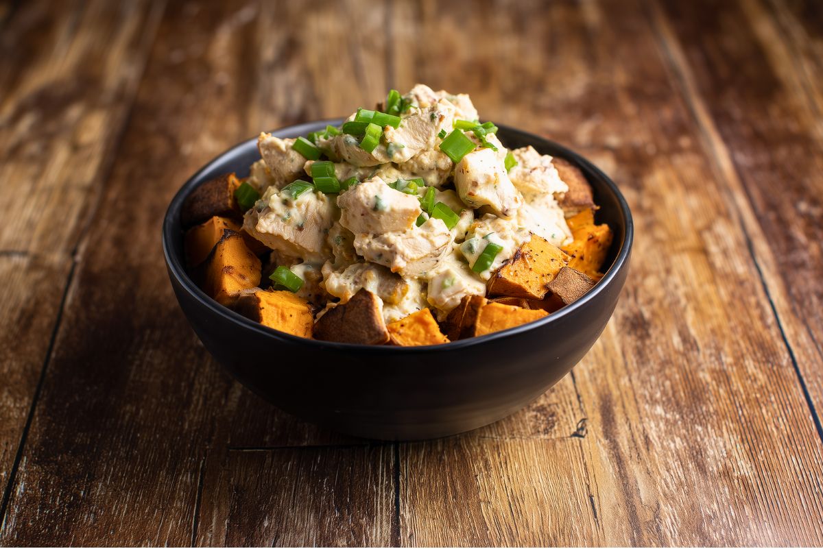Jalapeno Popper Chicken Sweet Potato Bowl with roasted sweet potato cubes topped with creamy chicken and fresh green onions, served in a black bowl on a rustic wood table.