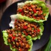 Three Kung Pao chicken lettuce wraps filled with glazed chicken, red bell peppers, peanuts, and green onions on a black plate over a rustic wood table.