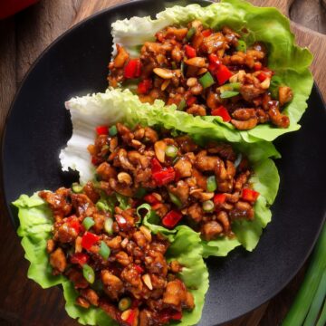 Three Kung Pao chicken lettuce wraps filled with glazed chicken, red bell peppers, peanuts, and green onions on a black plate over a rustic wood table.