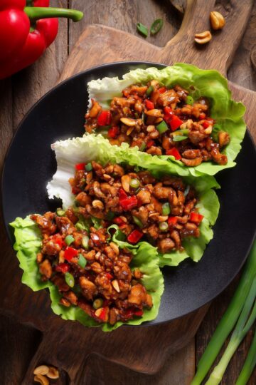 Three Kung Pao chicken lettuce wraps filled with glazed chicken, red bell peppers, peanuts, and green onions on a black plate over a rustic wood table.