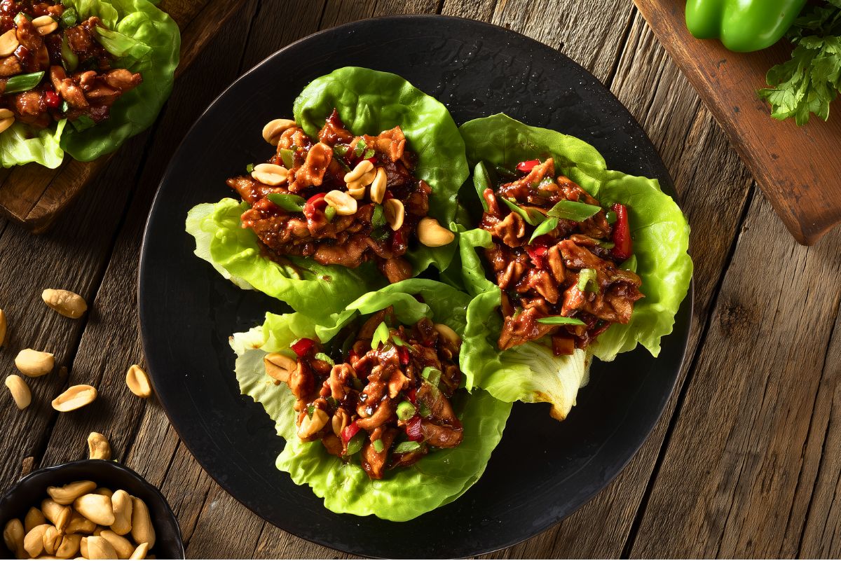 Overhead view of three Kung Pao chicken lettuce wraps with saucy chicken, red and green peppers, peanuts, and green onions on a black plate over rustic wood.
