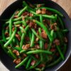 Close-up overhead view of maple bacon green beans with golden brown bacon pieces and a light maple glaze. Served on a matte black round plate over a rustic wooden table with a folded beige napkin.