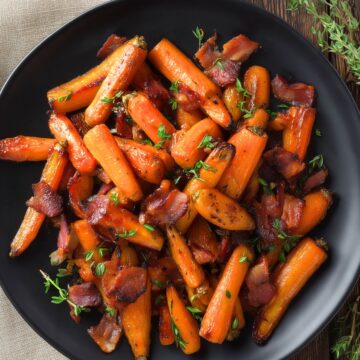 Overhead view of maple bacon roasted carrots glazed with maple syrup, mixed with crispy bacon pieces, and garnished with fresh thyme. Served on a matte black plate over dark rustic wood with a beige linen napkin.