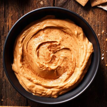 Overhead view of Maple Pumpkin Yogurt Dip in a black bowl on a dark rustic wooden table. The creamy orange dip has a cinnamon-dusted swirl, surrounded by graham crackers and red apple slices.