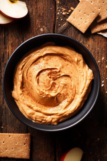 Overhead view of Maple Pumpkin Yogurt Dip in a black bowl on a dark rustic wooden table. The creamy orange dip has a cinnamon-dusted swirl, surrounded by graham crackers and red apple slices.