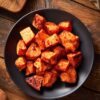 Overhead view of maple roasted sweet potatoes with golden orange centers, caramelized edges, and a glossy maple glaze. Served on a matte black plate over a rustic wooden table.