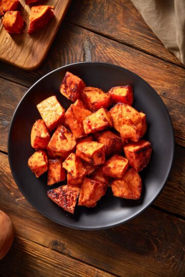 Overhead view of maple roasted sweet potatoes with golden orange centers, caramelized edges, and a glossy maple glaze. Served on a matte black plate over a rustic wooden table.