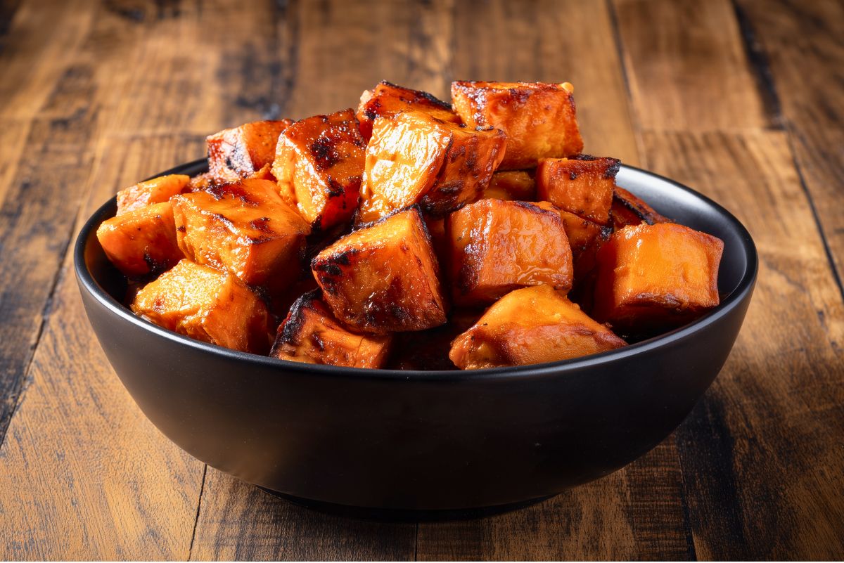 Close-up side view of maple roasted sweet potatoes piled high in a matte black bowl. The orange sweet potato cubes have glossy maple-coated surfaces and dark caramelized edges, with a rustic wooden table in the background.