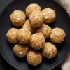 Overhead view of homemade Peanut Butter Banana Protein Balls stacked on a matte black plate. The golden brown, no-bake protein balls have visible rolled oats and a smooth, slightly textured surface. A beige cloth rests beside the plate.
