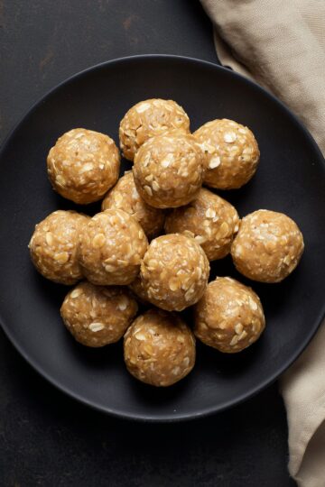 Overhead view of homemade Peanut Butter Banana Protein Balls stacked on a matte black plate. The golden brown, no-bake protein balls have visible rolled oats and a smooth, slightly textured surface. A beige cloth rests beside the plate.