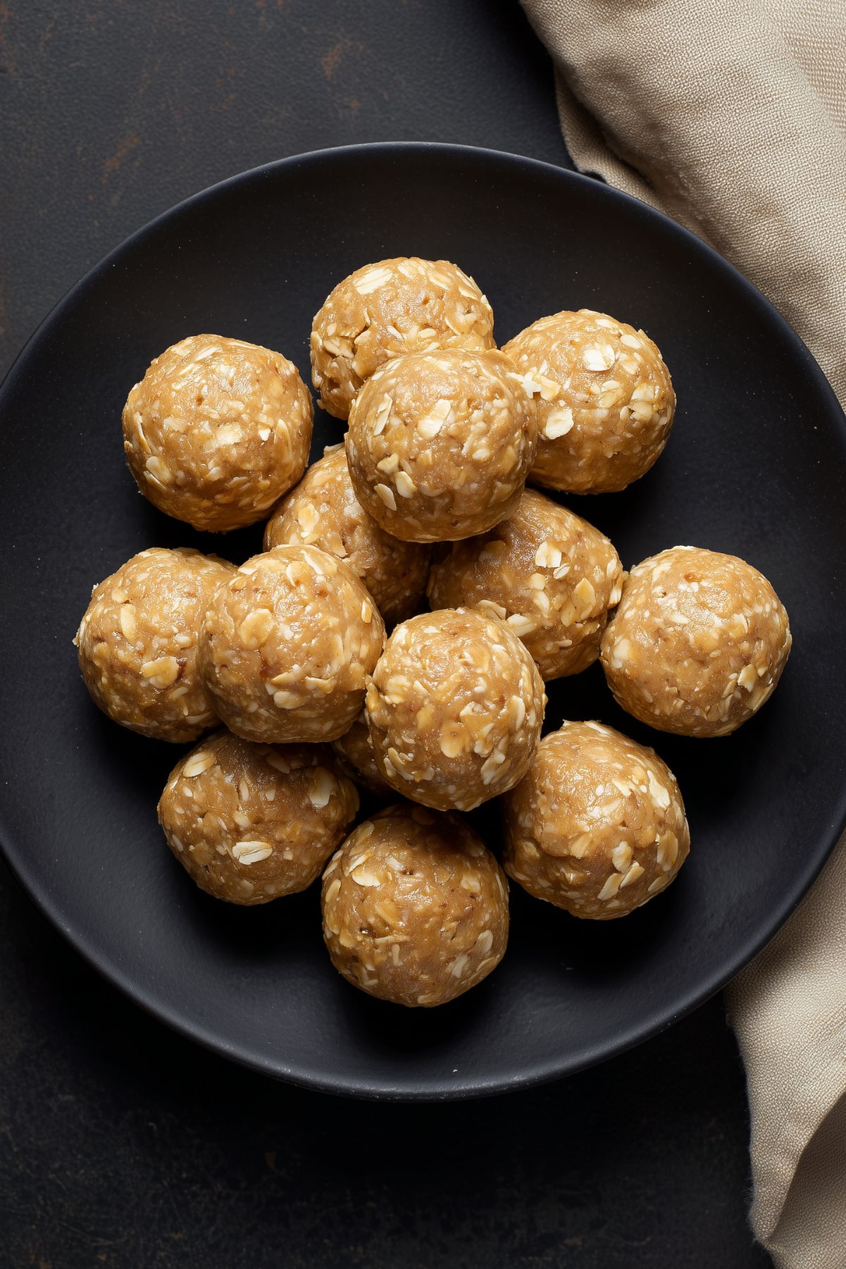 Overhead view of homemade Peanut Butter Banana Protein Balls stacked on a matte black plate. The golden brown, no-bake protein balls have visible rolled oats and a smooth, slightly textured surface. A beige cloth rests beside the plate.
