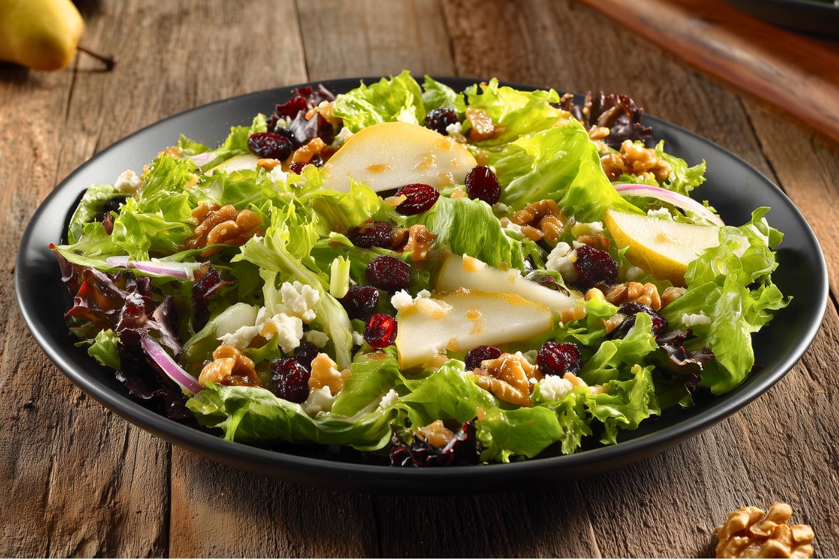 Pear Cranberry Walnut Salad with mixed greens, sliced pears, dried cranberries, walnuts, feta cheese, and red onion tossed with dressing on a black plate over a rustic wood table.