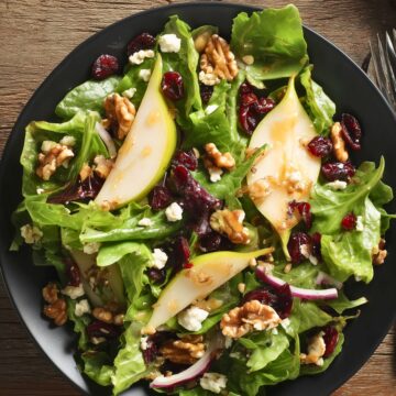 Overhead view of Pear Cranberry Walnut Salad with mixed greens, pear slices, dried cranberries, walnuts, feta cheese, red onion, and dressing served on a black plate.