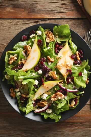 Overhead view of Pear Cranberry Walnut Salad with mixed greens, pear slices, dried cranberries, walnuts, feta cheese, red onion, and dressing served on a black plate.