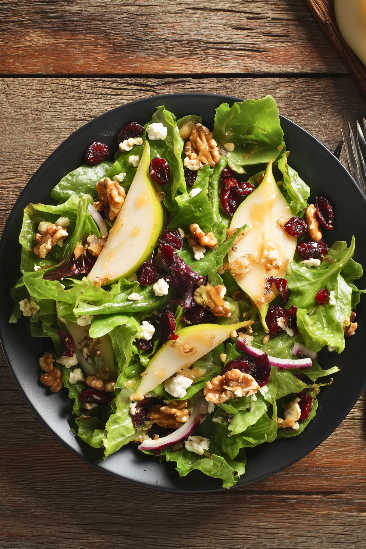 Overhead view of Pear Cranberry Walnut Salad with mixed greens, pear slices, dried cranberries, walnuts, feta cheese, red onion, and dressing served on a black plate.
