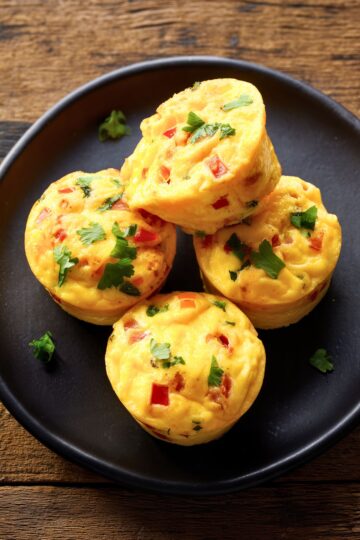 Close-up of four homemade Pico de Gallo Egg Bites with diced tomatoes, peppers, melted cheese, and fresh cilantro on a black plate.