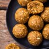 Overhead view of homemade Pumpkin Spice Protein Balls on a matte black plate over a rustic wooden table. Two protein balls have bites taken out, revealing a soft, chewy, oat-filled center.