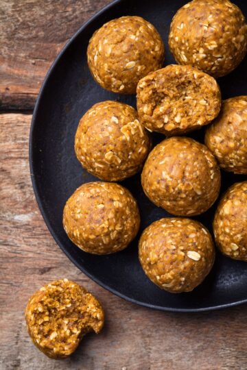 Overhead view of homemade Pumpkin Spice Protein Balls on a matte black plate over a rustic wooden table. Two protein balls have bites taken out, revealing a soft, chewy, oat-filled center.