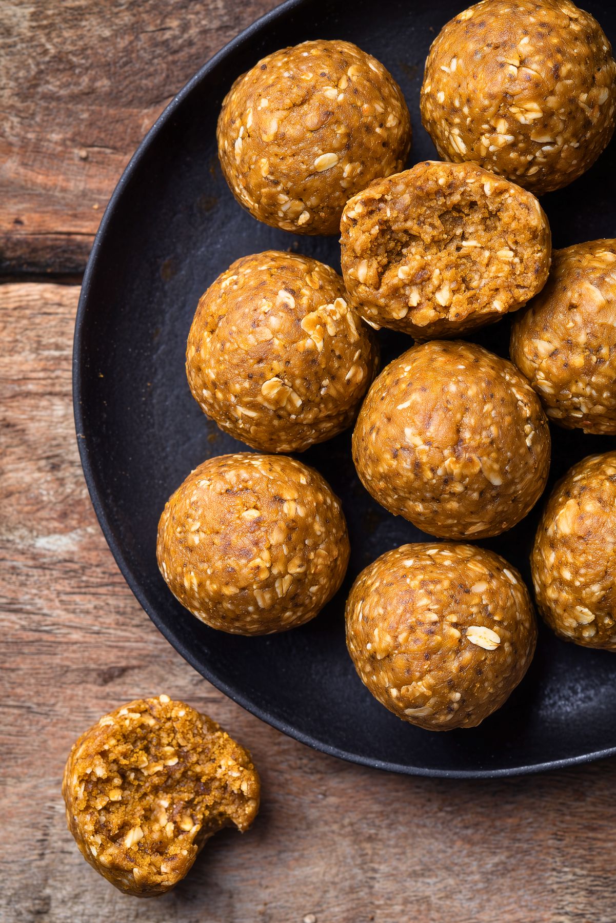 Overhead view of homemade Pumpkin Spice Protein Balls on a matte black plate over a rustic wooden table. Two protein balls have bites taken out, revealing a soft, chewy, oat-filled center.