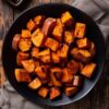 Pumpkin spice roasted sweet potatoes piled on a matte black plate, with glossy orange surfaces and dark caramelized edges. A baking sheet with additional roasted sweet potatoes sits in the upper left corner, with beige napkins on a rustic wooden table.