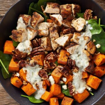 Close-up of a Rosemary Garlic Chicken Sweet Potato Bowl in a black bowl on a rustic wooden table. Golden brown chicken breast pieces, roasted sweet potato cubes, fresh spinach, toasted pecans, crumbled cheese, and creamy herb dressing.