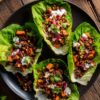 Overhead view of four Southwest beef and sweet potato lettuce wraps arranged on a black plate. Fresh lettuce cups are filled with seasoned ground beef, roasted sweet potatoes, yellow corn, red onion, tomatoes, cilantro, and creamy white dressing on a rustic wooden table.
