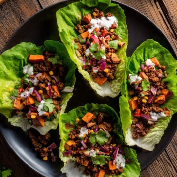 Overhead view of four Southwest beef and sweet potato lettuce wraps arranged on a black plate. Fresh lettuce cups are filled with seasoned ground beef, roasted sweet potatoes, yellow corn, red onion, tomatoes, cilantro, and creamy white dressing on a rustic wooden table.