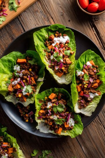 Overhead view of four Southwest beef and sweet potato lettuce wraps arranged on a black plate. Fresh lettuce cups are filled with seasoned ground beef, roasted sweet potatoes, yellow corn, red onion, tomatoes, cilantro, and creamy white dressing on a rustic wooden table.