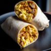 Close-up of two spicy chicken breakfast burrito halves stacked on a matte black plate. Soft flour tortillas are filled with seasoned chicken breast, fluffy scrambled eggs, melted cheese, and peppers.