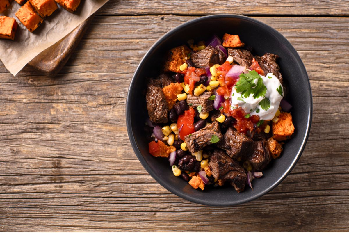 Steak burrito sweet potato bowl filled with seasoned steak chunks, roasted sweet potatoes, black beans, corn, red onion, salsa, and a dollop of sour cream topped with cilantro. Served in a matte black bowl on a rustic wooden table with extra roasted sweet potatoes nearby.
