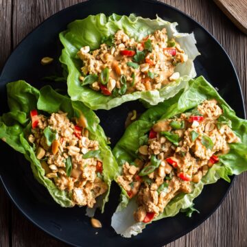 Three Thai peanut chicken lettuce wraps on a black plate, filled with ground chicken, creamy peanut sauce, red peppers, chopped peanuts, green onions, and cilantro.