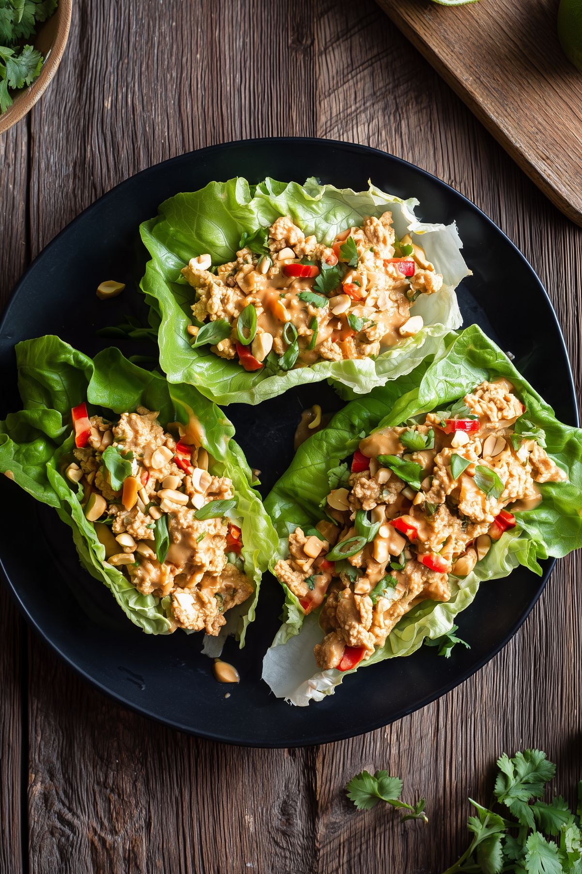 Three Thai peanut chicken lettuce wraps on a black plate, filled with ground chicken, creamy peanut sauce, red peppers, chopped peanuts, green onions, and cilantro.