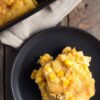 A serving of Thanksgiving Corn Casserole on a matte black plate, showing a moist, tender cornbread-like texture with yellow corn kernels and golden edges. A partially served baking dish rests on a beige linen cloth over a rustic wooden table.