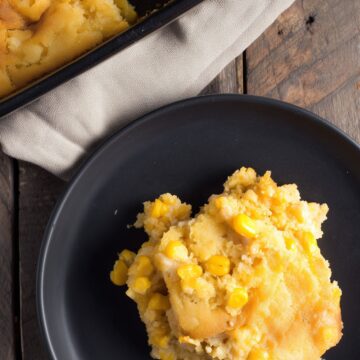 A serving of Thanksgiving Corn Casserole on a matte black plate, showing a moist, tender cornbread-like texture with yellow corn kernels and golden edges. A partially served baking dish rests on a beige linen cloth over a rustic wooden table.
