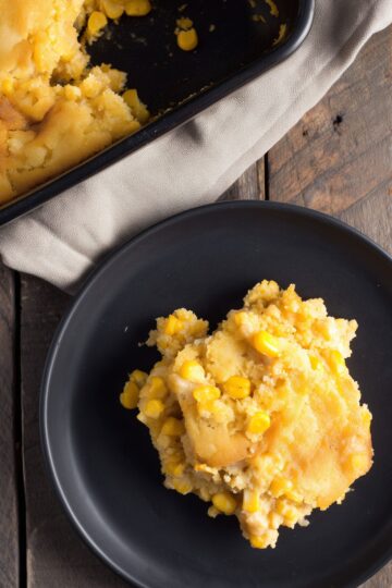 A serving of Thanksgiving Corn Casserole on a matte black plate, showing a moist, tender cornbread-like texture with yellow corn kernels and golden edges. A partially served baking dish rests on a beige linen cloth over a rustic wooden table.