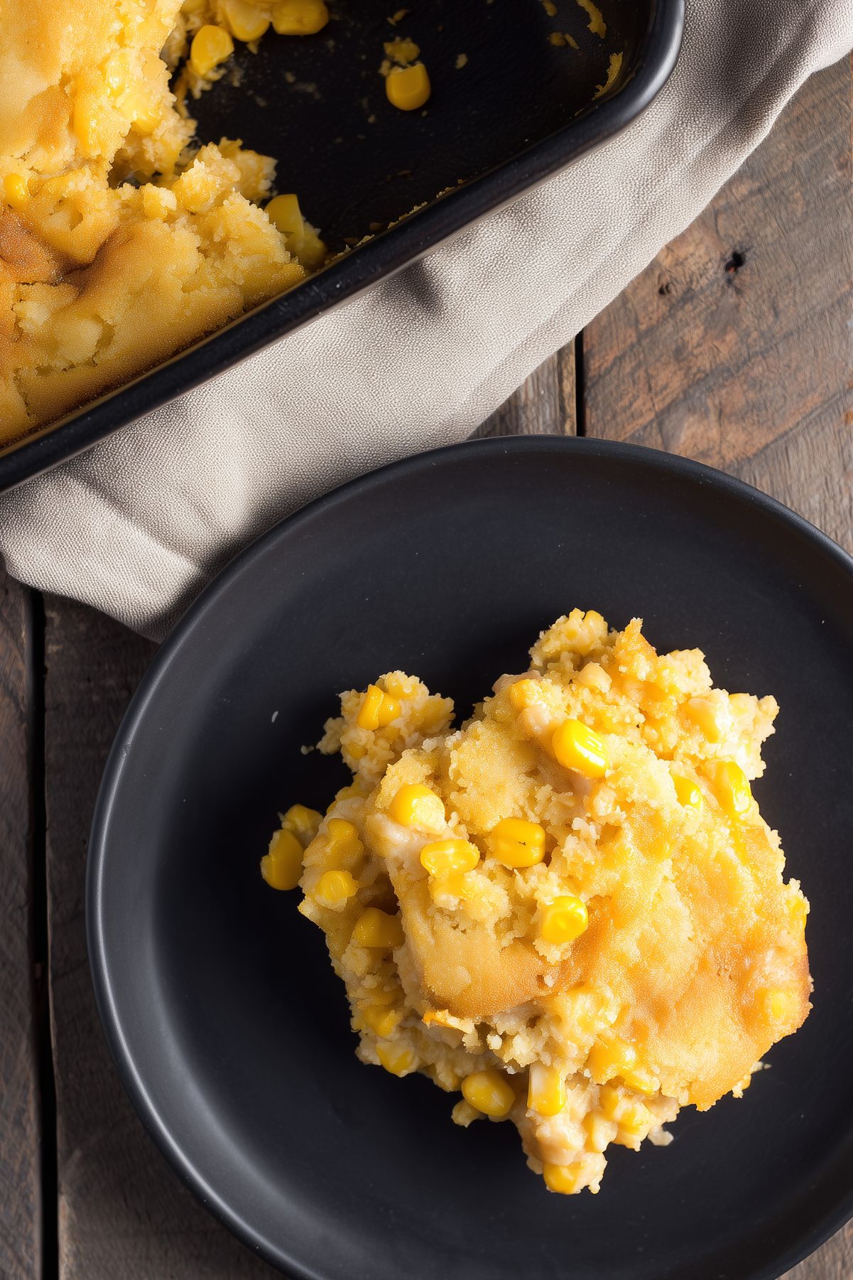 A serving of Thanksgiving Corn Casserole on a matte black plate, showing a moist, tender cornbread-like texture with yellow corn kernels and golden edges. A partially served baking dish rests on a beige linen cloth over a rustic wooden table.