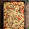 Overhead view of Tomato Parmesan Spaghetti Squash Casserole baked in a rectangular pan, topped with melted Parmesan cheese, chopped tomatoes, basil, and herbs on a rustic wood table.