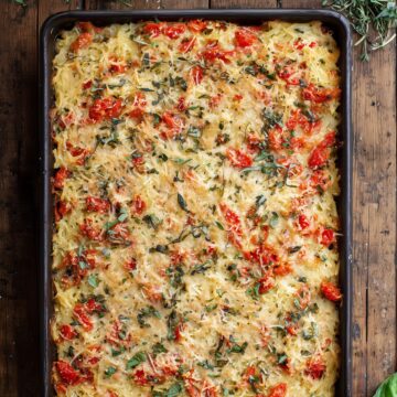Overhead view of Tomato Parmesan Spaghetti Squash Casserole baked in a rectangular pan, topped with melted Parmesan cheese, chopped tomatoes, basil, and herbs on a rustic wood table.
